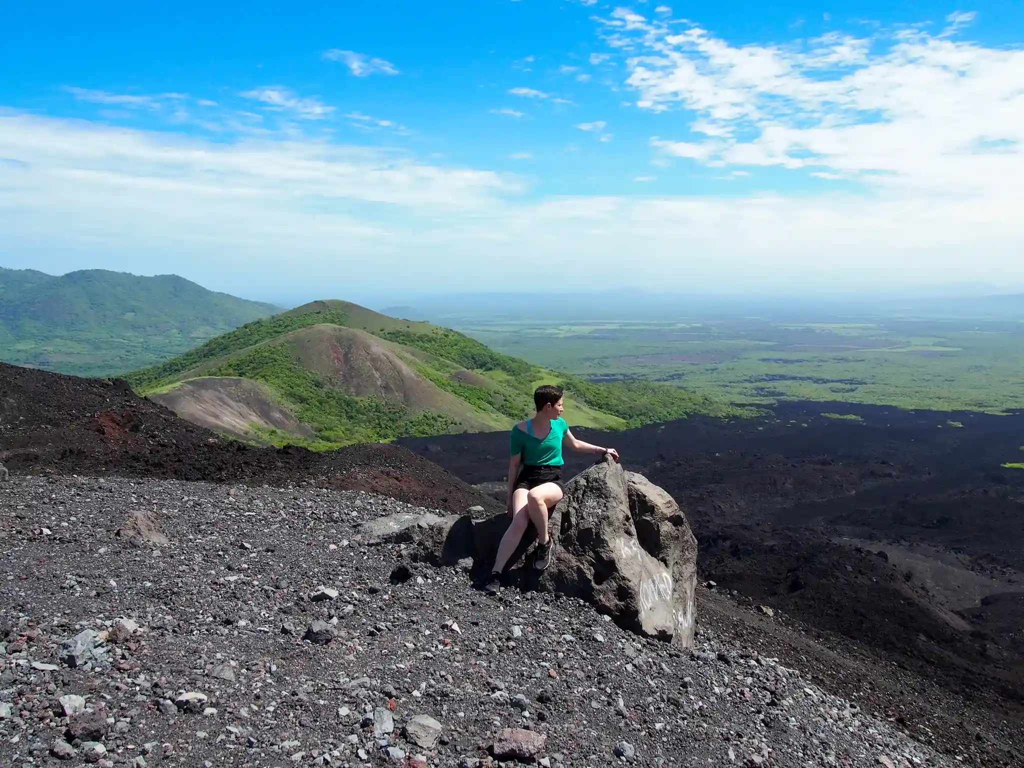 Imagen del paisaje desde la cima de una montaña, cielo despejado, montañas a los lados y rocas volcánicas en el suelo, siendo observado por una mujer sentada en una roca.